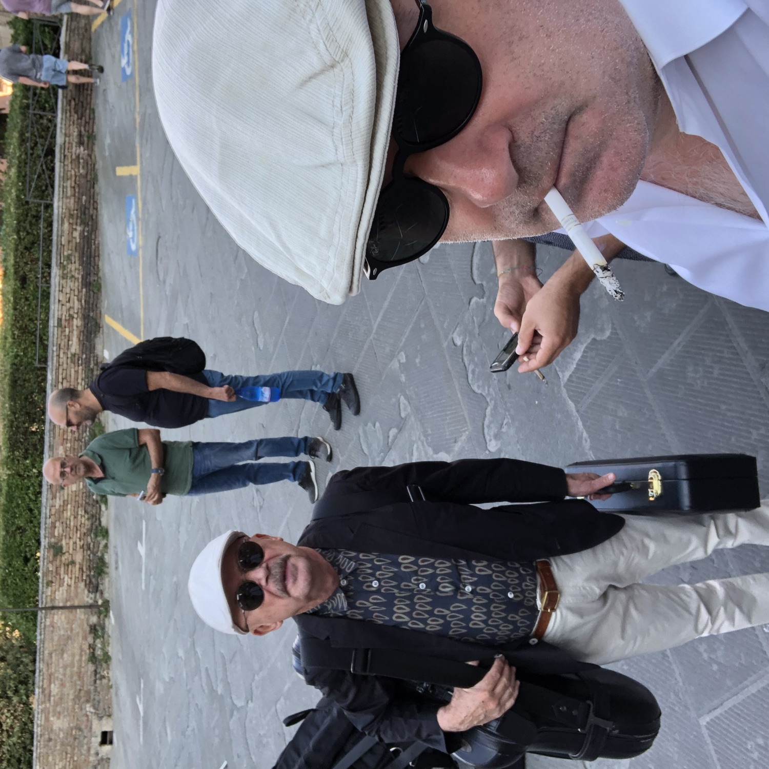 Michael Gassmann in a flat cap and sunglasses at an open-air festival; a fellow musician with an instrument case behind